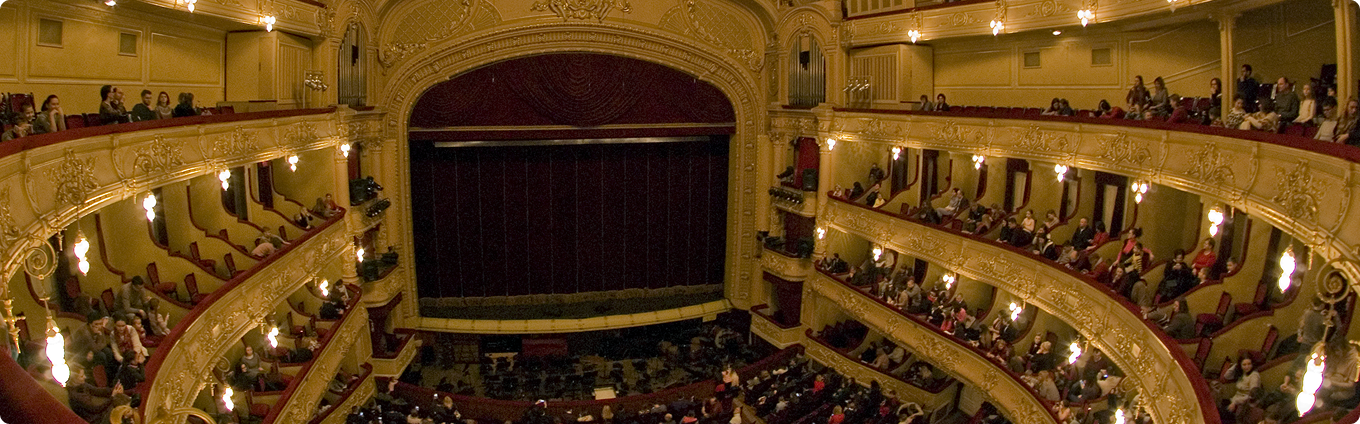 Панорамний вид внутрішнього залу театру з балконами, партером і сценою з опущеним занавісом.